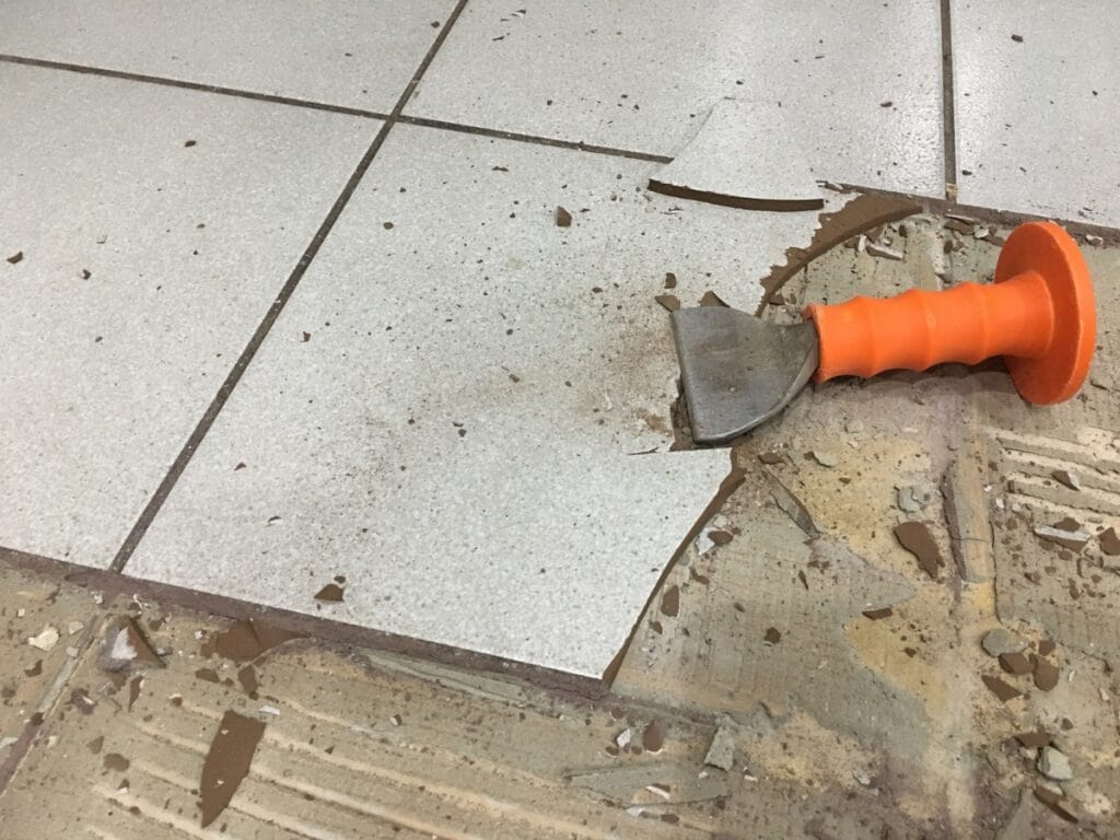 A chisel with an orange handle rests on a floor amid partially removed tiles and broken tile pieces—evidence of ongoing flooring removal in Marietta.