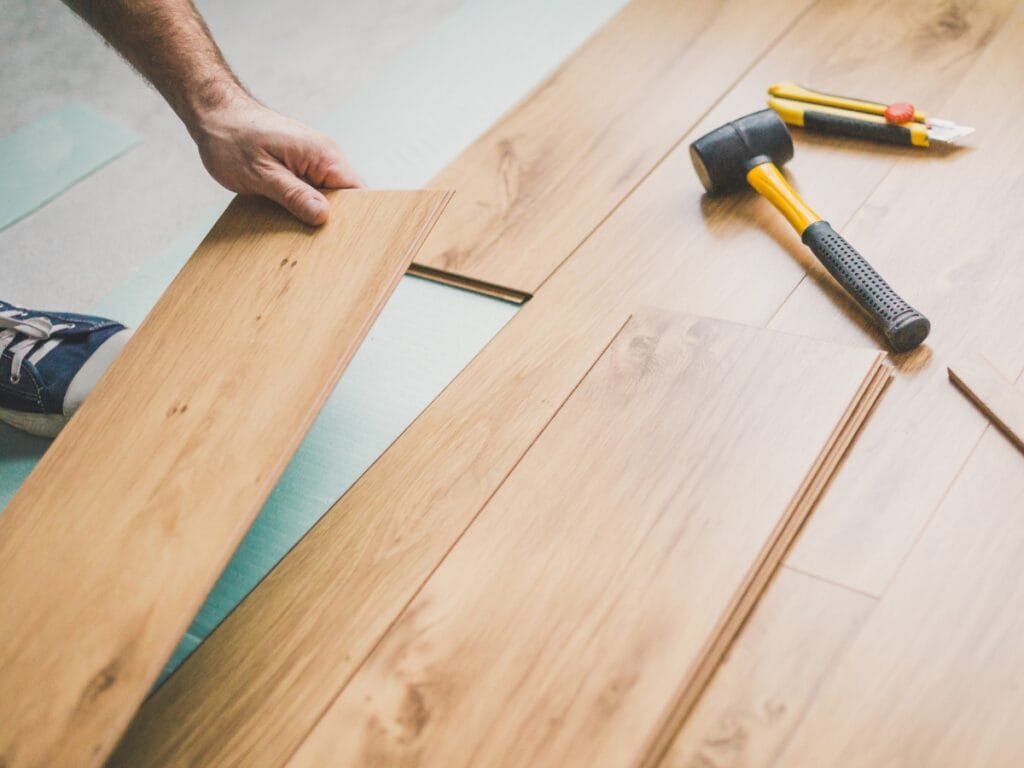 A person installs wooden laminate flooring, fitting a plank into place. Tools for flooring repair Marietta, including a rubber mallet, utility knife, and tape measure, are on the floor nearby.