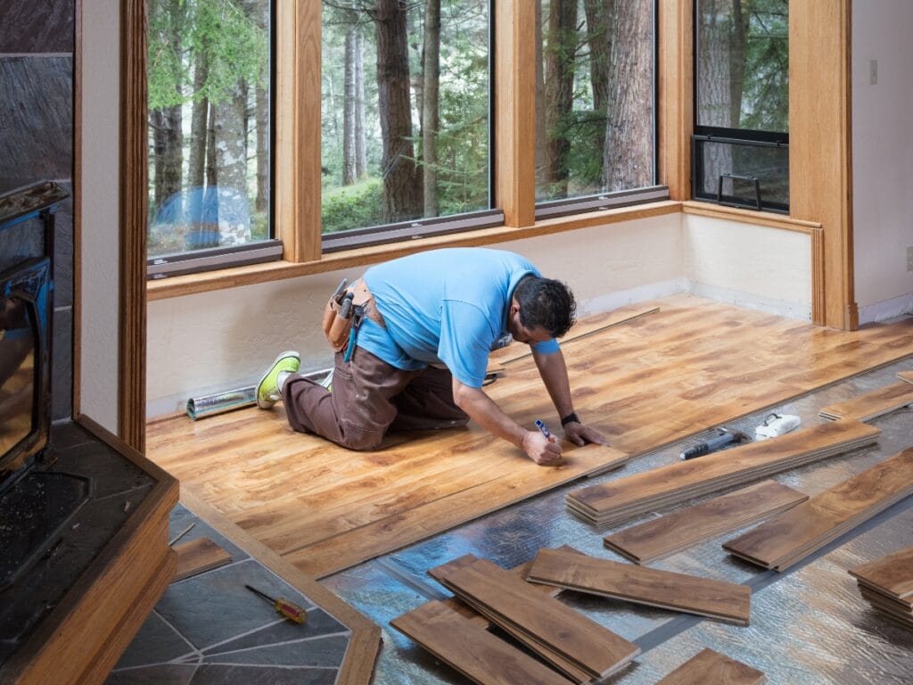 A person kneels on the floor, measuring and installing wood flooring in a room with large windows overlooking a forest, showcasing expert flooring repair Marietta craftsmanship.