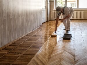 A worker wearing protective gear applies a clear finish to a wooden parquet floor in a bright, empty room with a paint roller, showcasing expert flooring repair Marietta services.