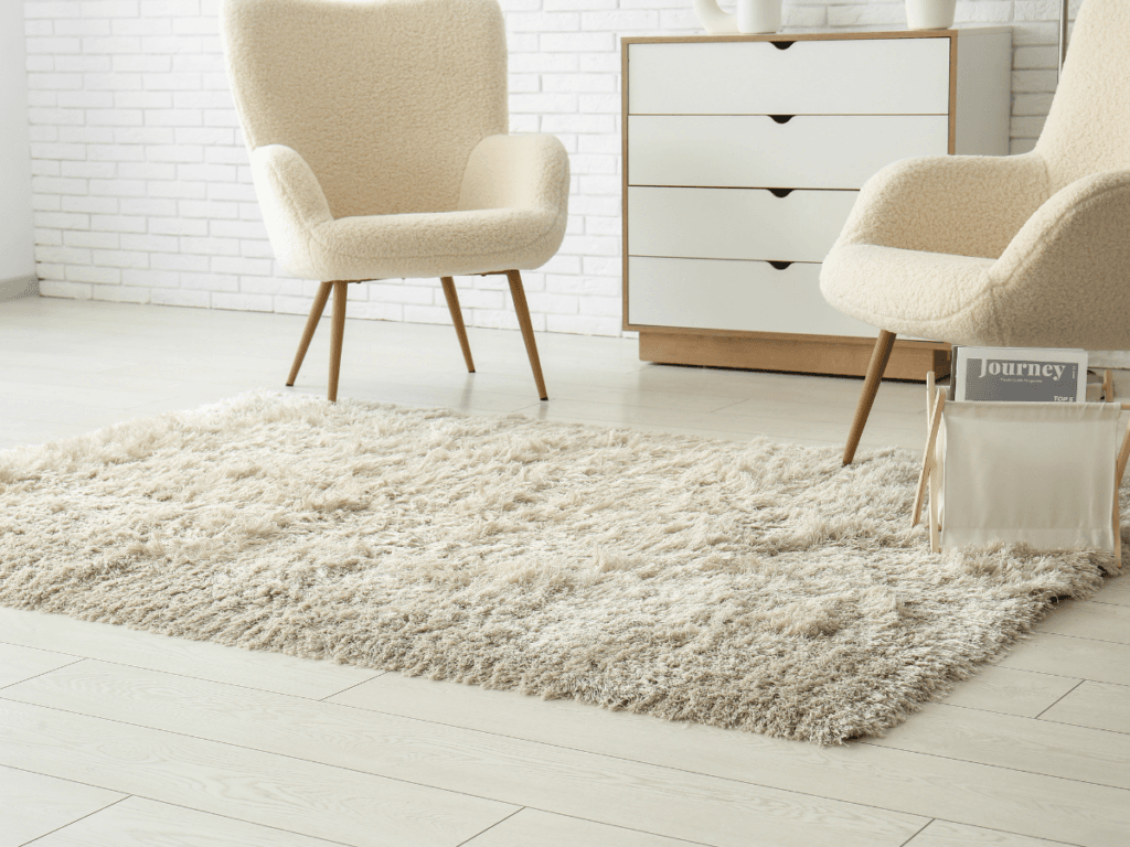 A modern living room with two beige armchairs, a fluffy beige rug, a white dresser with wood accents, and a magazine rack on light wood flooring offers inspiration for any carpet flooring padding guide.