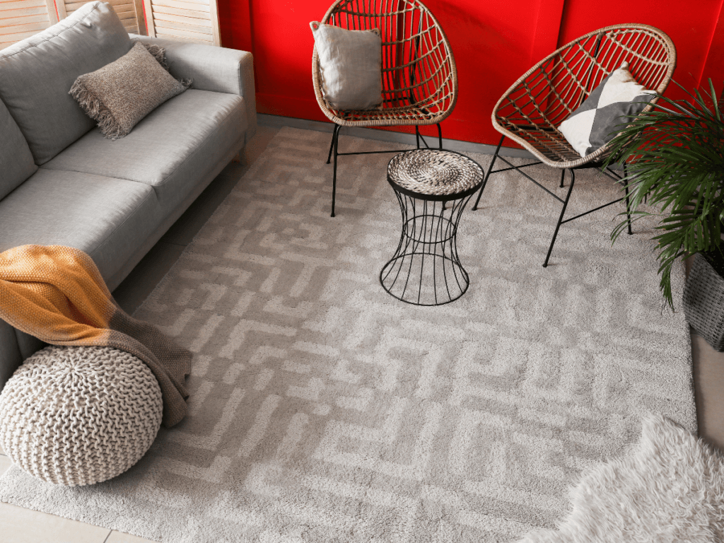 A modern living room with a gray sofa, two woven chairs, a wire-frame side table, a round knit pouf, and a patterned light gray rug atop plush carpet flooring&mdash;perfect for comfort with the right padding guide.