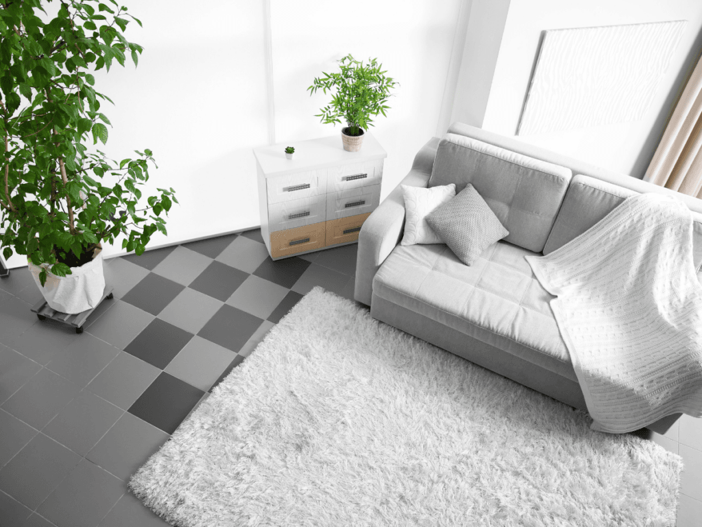 A modern living room with a gray sofa, white blanket, two pillows, white rug atop checkered tile floor, potted plants, a white chest of drawers, and decor inspired by our carpet flooring padding guide.
