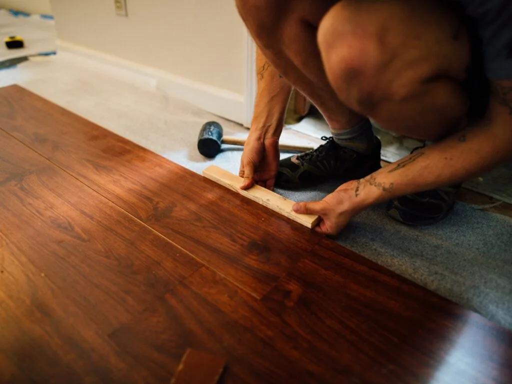 Person installing hardwood flooring, using a wooden block to fit planks together. A rubber mallet and partially finished floor are visible, making this scene ideal for a Luxury Vinyl Plank Click Lock Guide illustration.