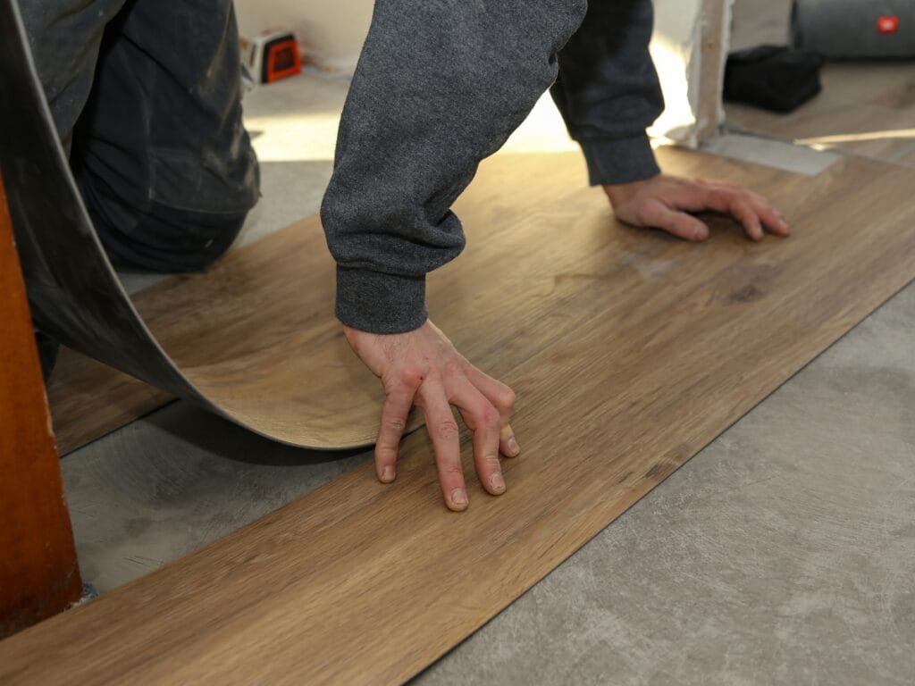 A person installs a sheet of wood-look vinyl flooring onto a concrete subfloor, pressing the material down with their hands, following tips from the Luxury Vinyl Plank Click Lock Guide for best results.