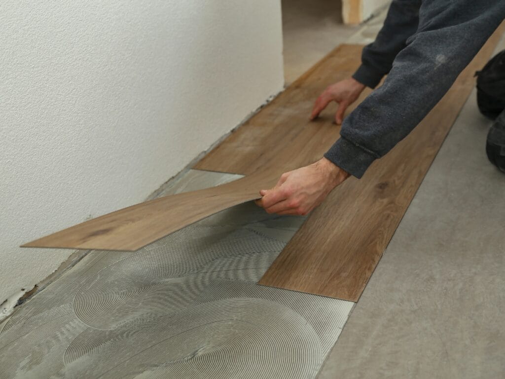 A person installs brown luxury vinyl plank flooring, placing the planks onto adhesive spread on a concrete subfloor near a white wall, following a Luxury Vinyl Plank Click Lock Guide.