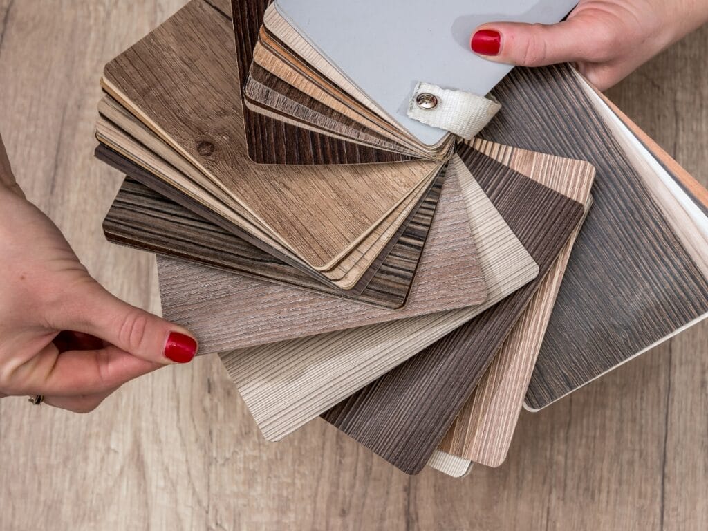 Hands holding a fan of wood laminate samples in various shades and grain patterns, displayed over a wooden surface—perfect inspiration for your next project or Luxury Vinyl Plank Click Lock Guide.