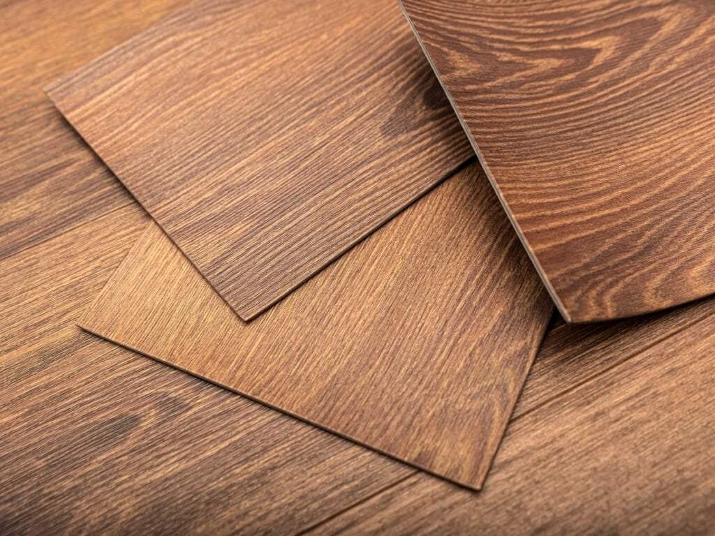 Three rectangular sheets of wood-patterned laminate flooring are shown arranged in an overlapping manner on top of a matching wood-patterned floor, illustrating options in the Luxury Vinyl Plank Click Lock Guide.
