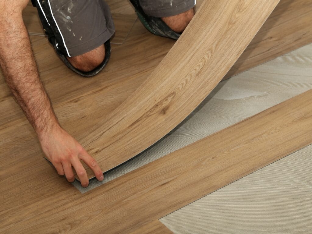 A person installs a light brown wood-look vinyl plank floor, positioning one plank over adhesive strips on the subfloor using tips from the Luxury Vinyl Plank Click Lock Guide.