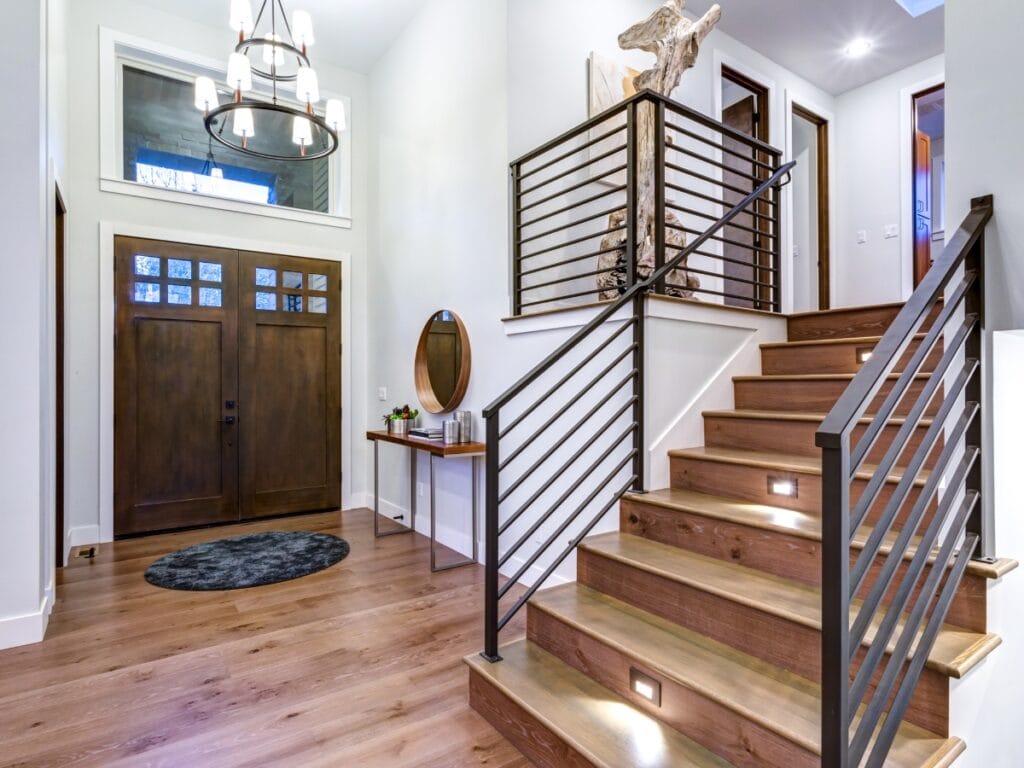 Modern entryway with wooden stairs, metal railing, double front doors, a chandelier, round mirror on a console table, and hardwood flooring&mdash;perfect inspiration for residential entryway ideas.