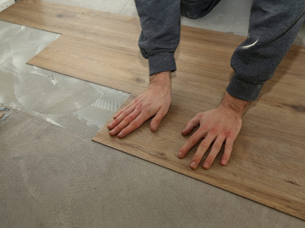 A person installs wood-look vinyl flooring, pressing a plank onto adhesive spread on a concrete subfloor&mdash;an inspiring choice for those seeking residential entryway ideas.