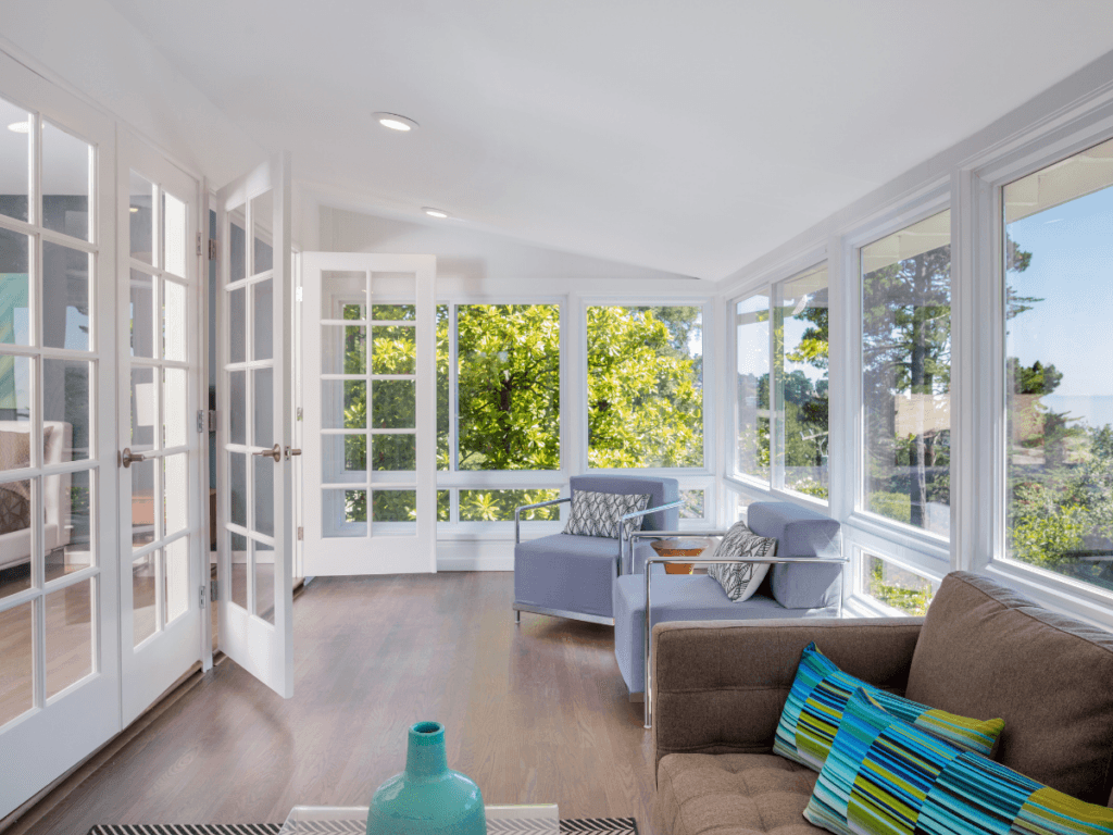 Bright sunroom with large windows, stylish sunroom flooring, a brown sofa, blue armchair, colorful pillows, and a view of green trees outside. Glass doors open to an adjacent room.