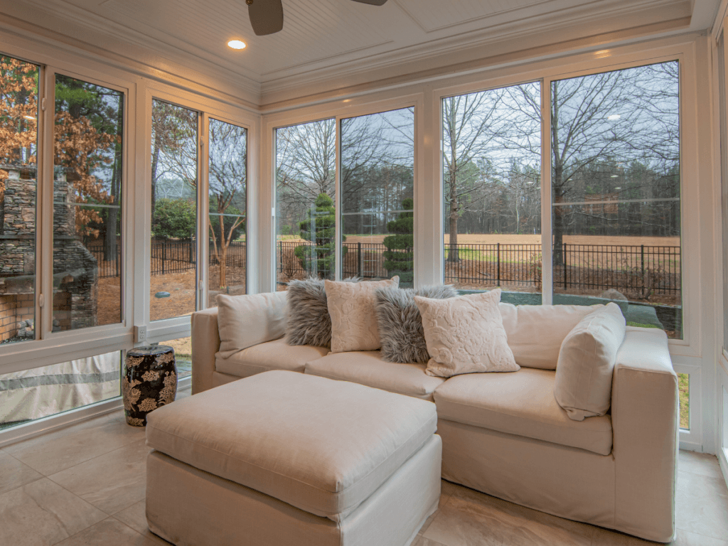 A bright sunroom with large windows, a white sectional sofa with decorative pillows, stylish sunroom flooring, and a view of a fenced backyard with trees.