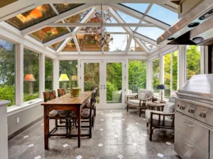 Sunroom kitchen with glass ceiling and walls, a wooden dining table with chairs, armchairs, stainless steel grill, elegant sunroom flooring, and beautiful views of greenery outside.