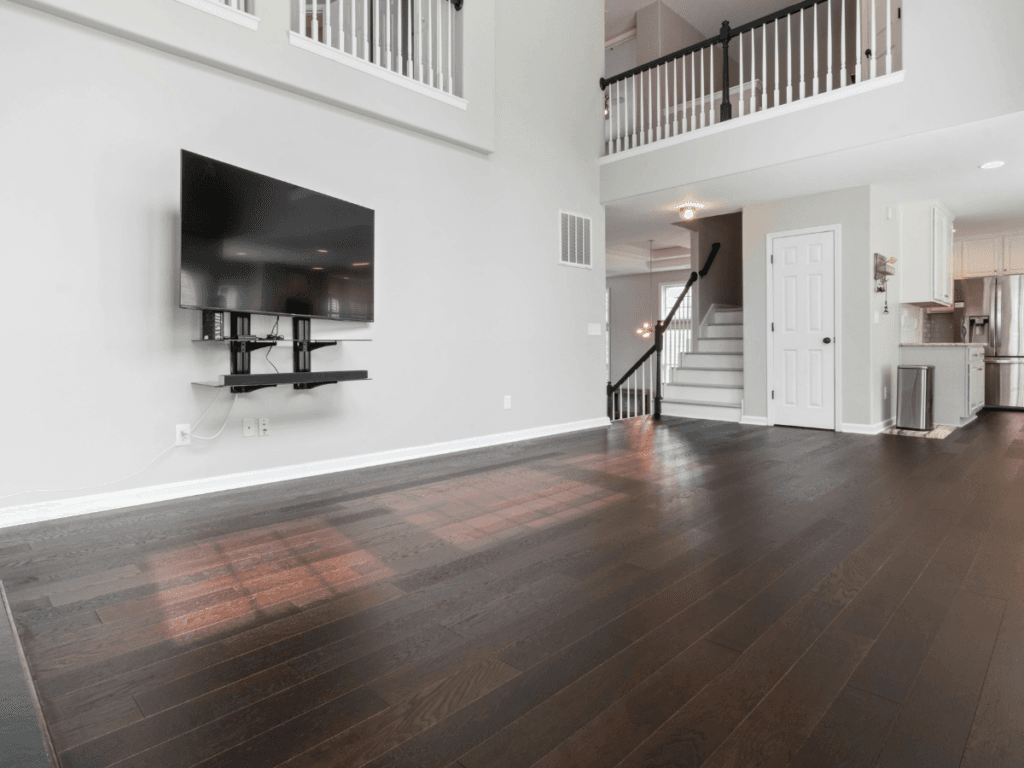 Spacious living room with Marietta allergy flooring, dark wood accents, wall-mounted TV, open stairway, and a view of a modern kitchen with white cabinets in the background.