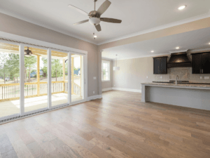 Spacious, empty living area with Marietta allergy flooring, hardwood floors, ceiling fan, and large sliding glass doors to a covered patio; kitchen with island and dark cabinets in the background.