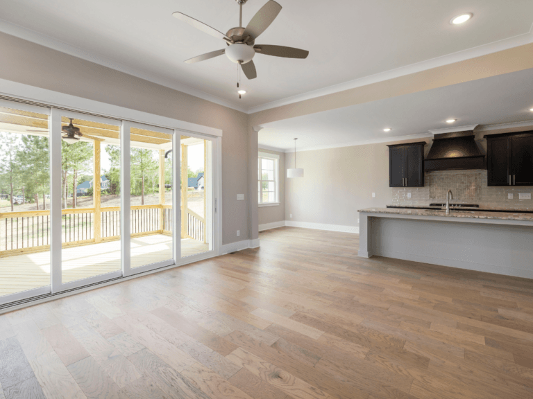 Spacious, empty living area with Marietta allergy flooring, hardwood floors, ceiling fan, and large sliding glass doors to a covered patio; kitchen with island and dark cabinets in the background.
