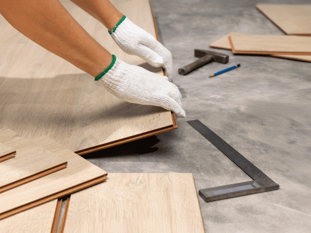 A person wearing white gloves installs Marietta pet flooring, laying wooden laminate on a concrete surface, with tools like a square ruler, pencil, and hammer nearby.