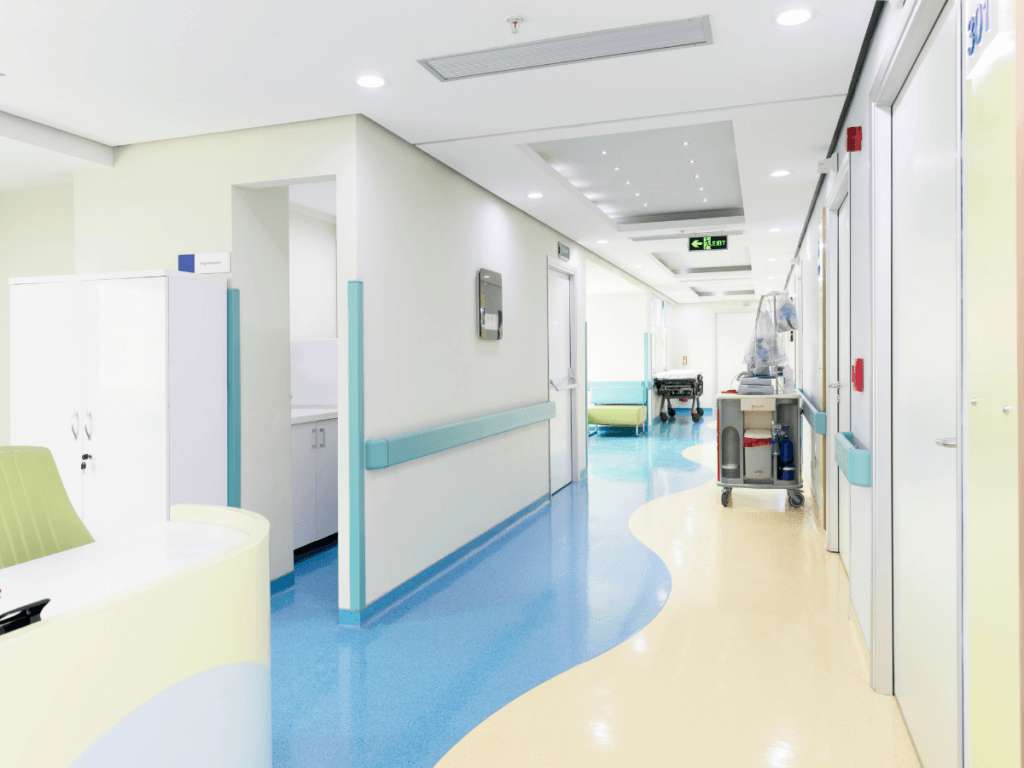 A clean, brightly lit hospital hallway with blue and yellow school flooring, a medical equipment cart, and open doors leading to patient rooms.