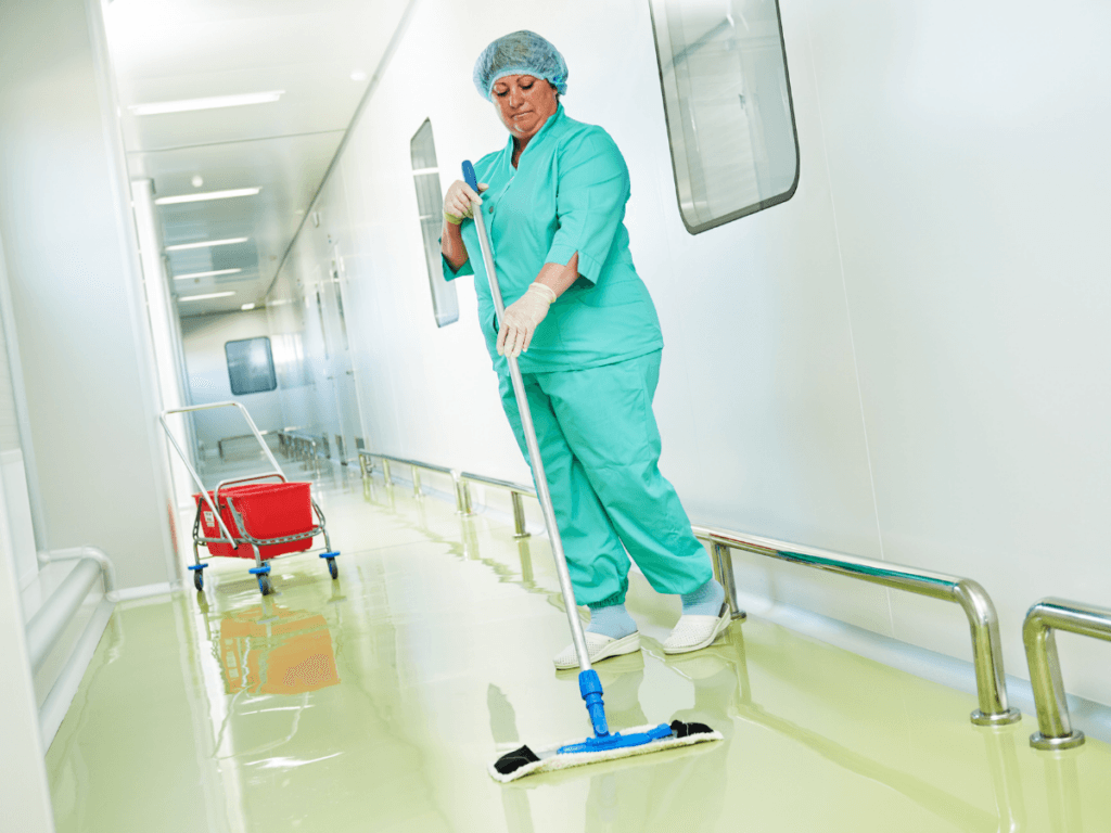 A person in medical scrubs and gloves mops the floor of a bright, clean hallway with a mop bucket nearby, ensuring the school flooring remains spotless and hygienic.
