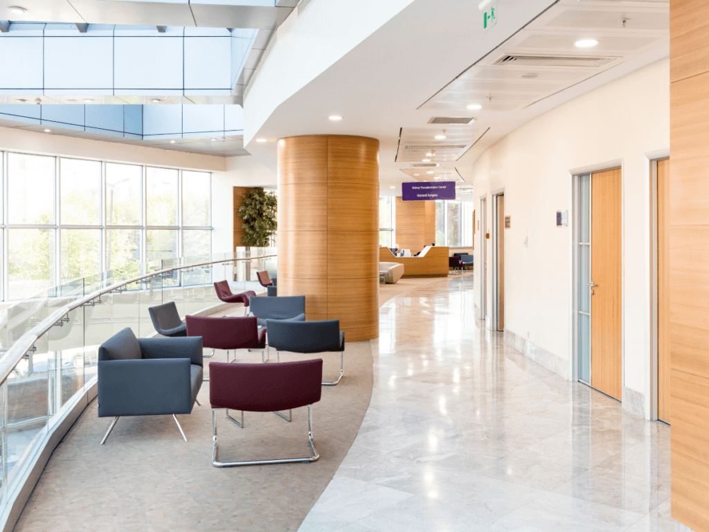 Modern, bright hospital waiting area with armchairs, curved glass railing, wood-paneled columns, and large windows letting in natural light. Durable school flooring adds a clean, resilient touch to the space as several doors line the hallway.