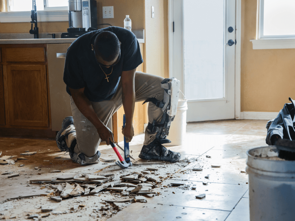 A person wearing knee pads is engaged in tile removal, using tools to take out broken tiles from a floor in a kitchen or dining area with cabinets and sunlight streaming through a window.