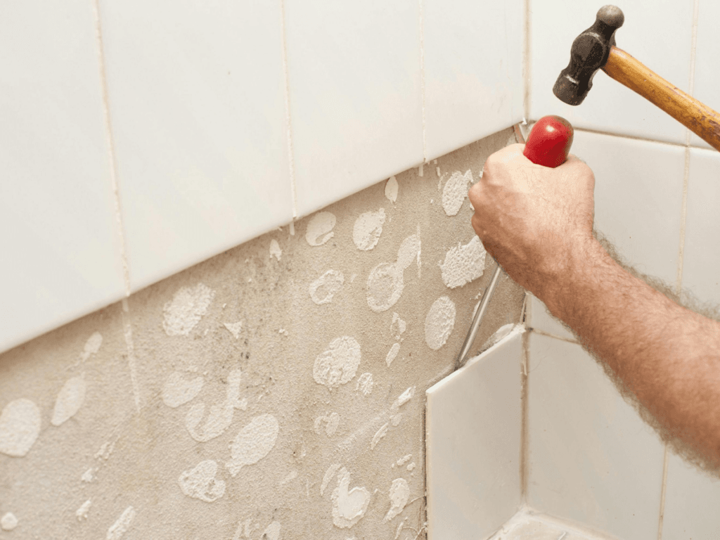 A person performs tile removal by using a hammer and chisel to remove white ceramic tiles from a wall, revealing adhesive and the unfinished surface underneath.
