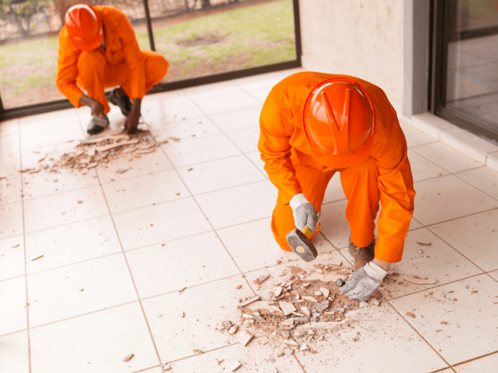 Two workers in orange uniforms and helmets perform tile removal, using hand tools and gloves to clear broken tiles and debris from a tiled floor inside a building.