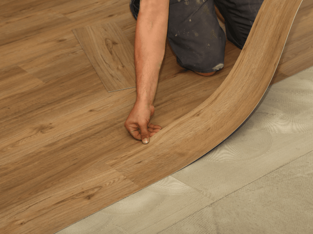 A person installing wood-look vinyl flooring, carefully bending a plank to fit into place over adhesive on the subfloor&mdash;demonstrating a technique that&rsquo;s useful for both new installations and flooring repair, such as fixing LVP scratches.