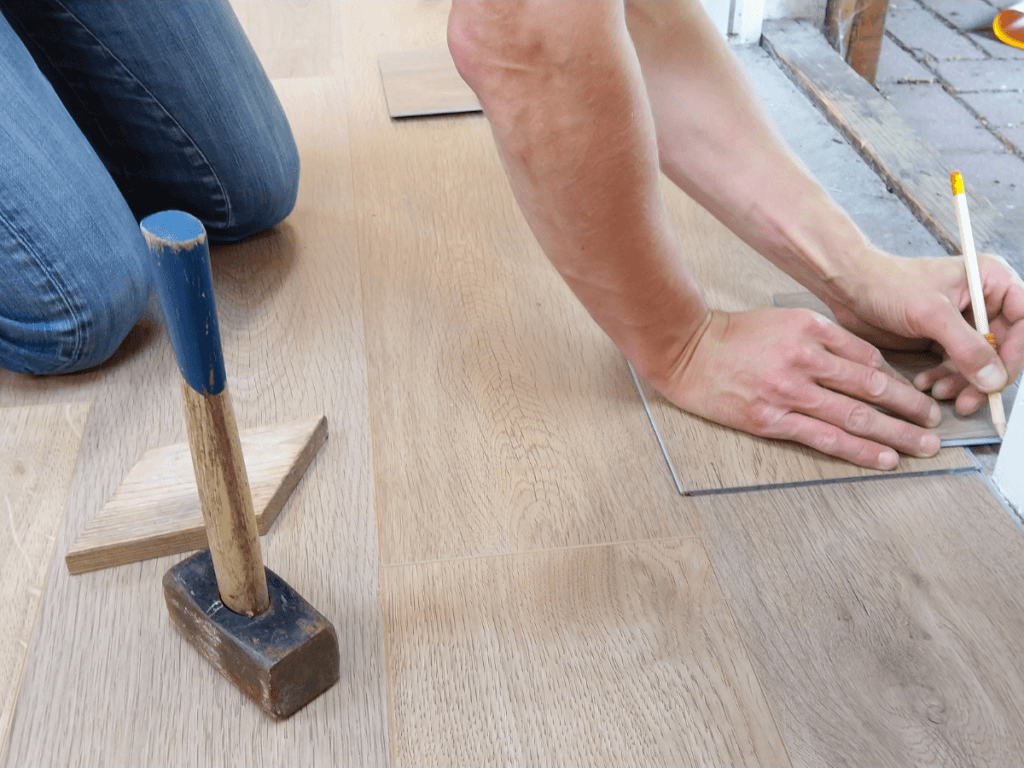Person measuring and marking a piece of laminate flooring for cutting, with a hammer and wooden block nearby on the floor&mdash;an essential step in any flooring repair, especially when addressing LVP scratches or replacing damaged planks.