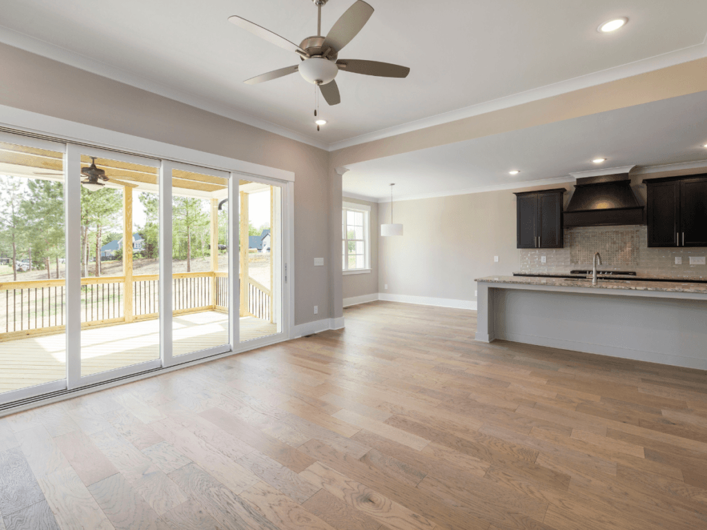 Empty open-plan living area with wood floors, ceiling fan, and large glass doors leading to a covered patio; adjacent kitchen with dark cabinets and a breakfast bar. The space highlights beautiful flooring and has been thoughtfully maintained, minimizing the need for flooring repair or concerns about LVP scratches.